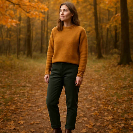 En kvinna i bruna chelsea boots dam brun, klädd i mörkgrön chinos och en mjuk ockerfärgad ulltröja, stående i en skogsglänta under höstlöven. Alt: brun chelsea boot dam stil färgkombination skog stad